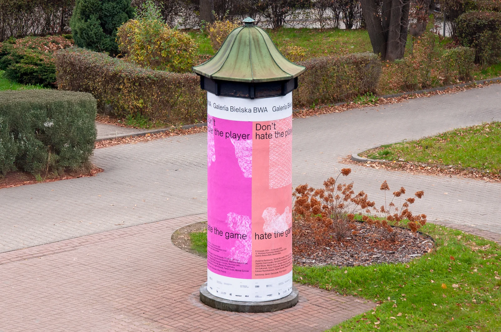 Słup ogłoszeniowy w parku oklejony dużymi różowymi i brzoskwiniowymi plakatami wystawy w Galerii Bielskiej BWA.