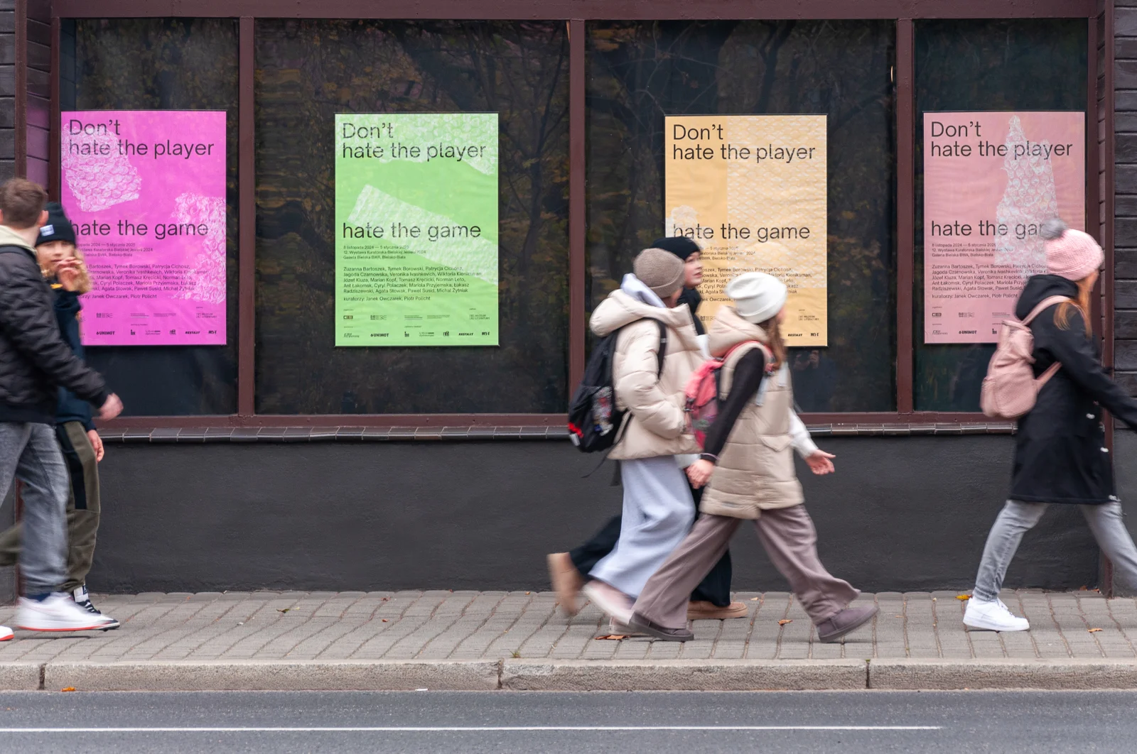 Przechodnie mijają witrynę galerii, w której wyeksponowano cztery kolorowe plakaty wystawy w kolorach różowym, zielonym, żółtym i beżowym.