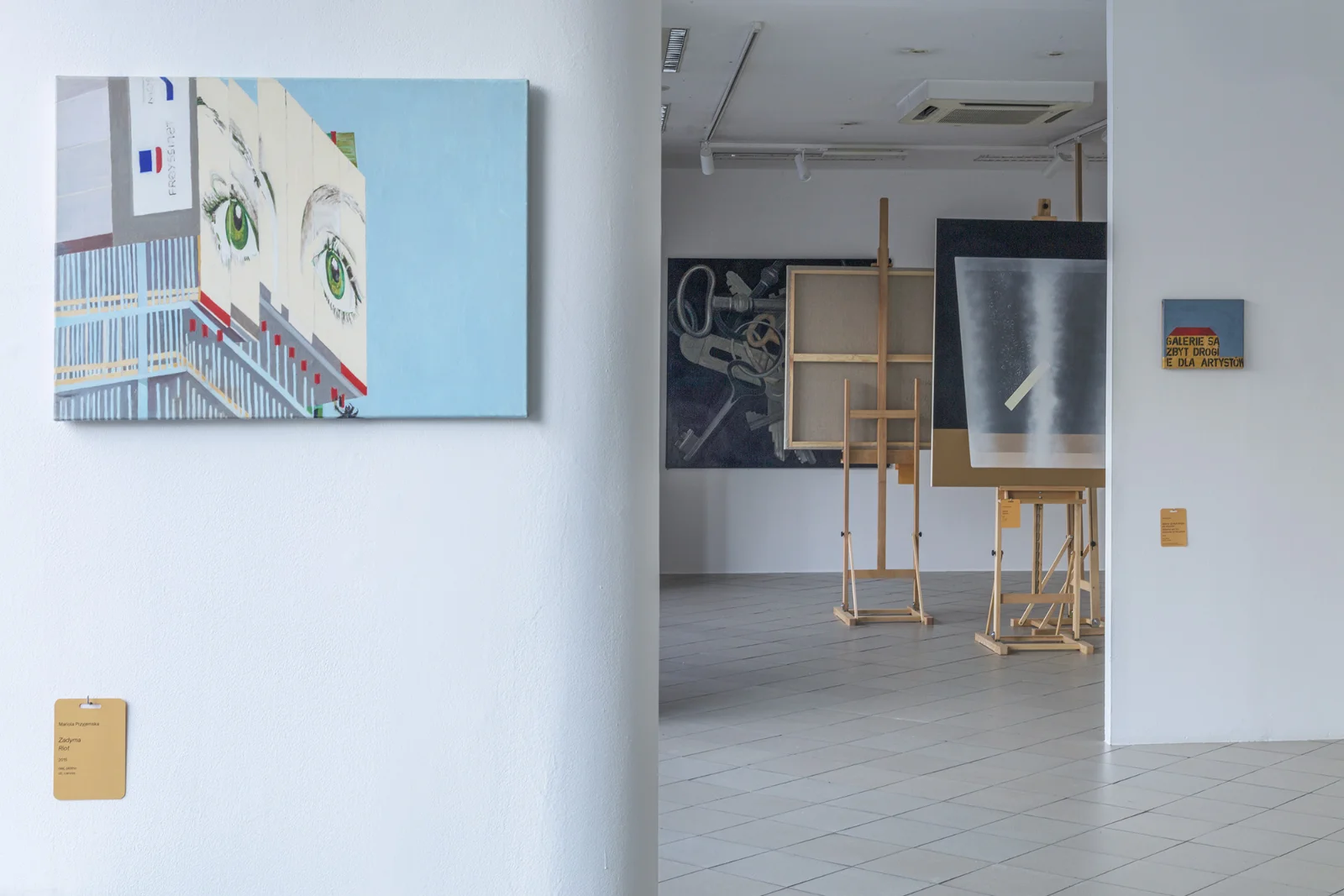 Widok na galerię przez przejście; na pierwszym planie obraz przedstawiający duże zielone oczy, w tle sztalugi i różne dzieła sztuki.