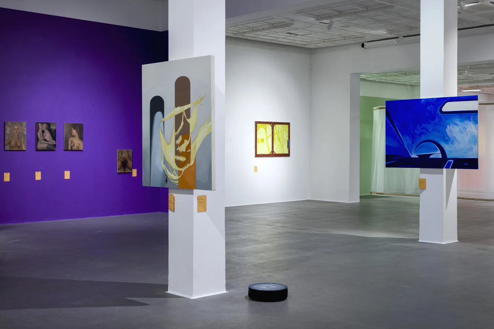 Szeroki widok wnętrza galerii z fioletową ścianą po lewej stronie i wielkoformatowymi obrazami zamontowanymi na białych filarach w otwartej hali.