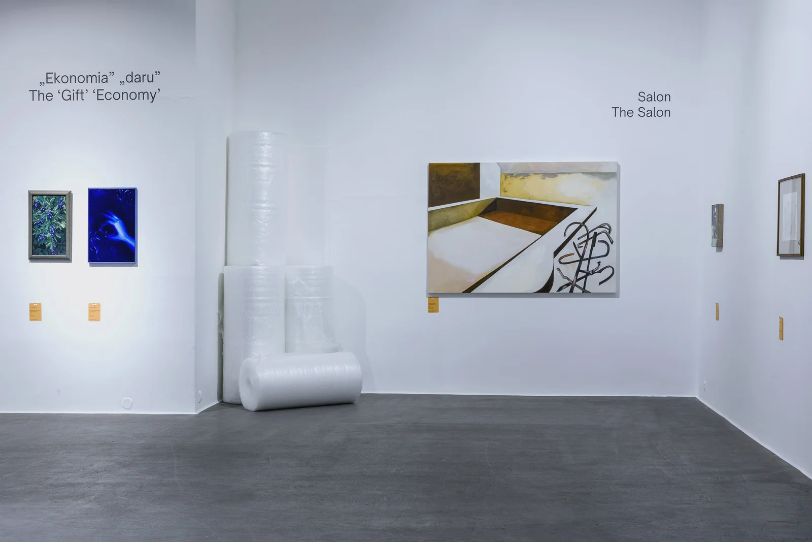 Narożnik galerii z białymi ścianami; rolki folii bąbelkowej stoją w kącie obok obrazów pod napisami „Ekonomia daru” i „Salon”.