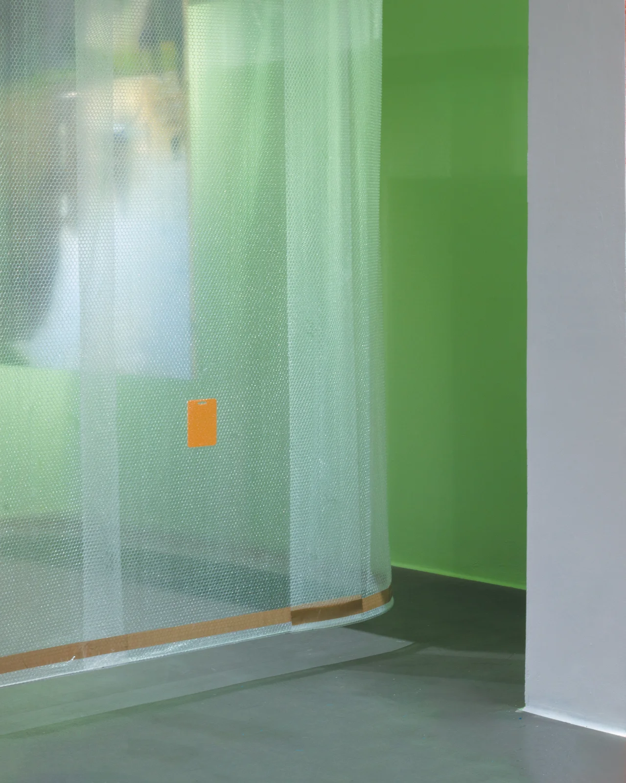 Zbliżenie na półprzezroczystą kurtynę z folii bąbelkowej wiszącą w galerii z żółtą zawieszką z tytułem obrazu na tle zielonej ściany.