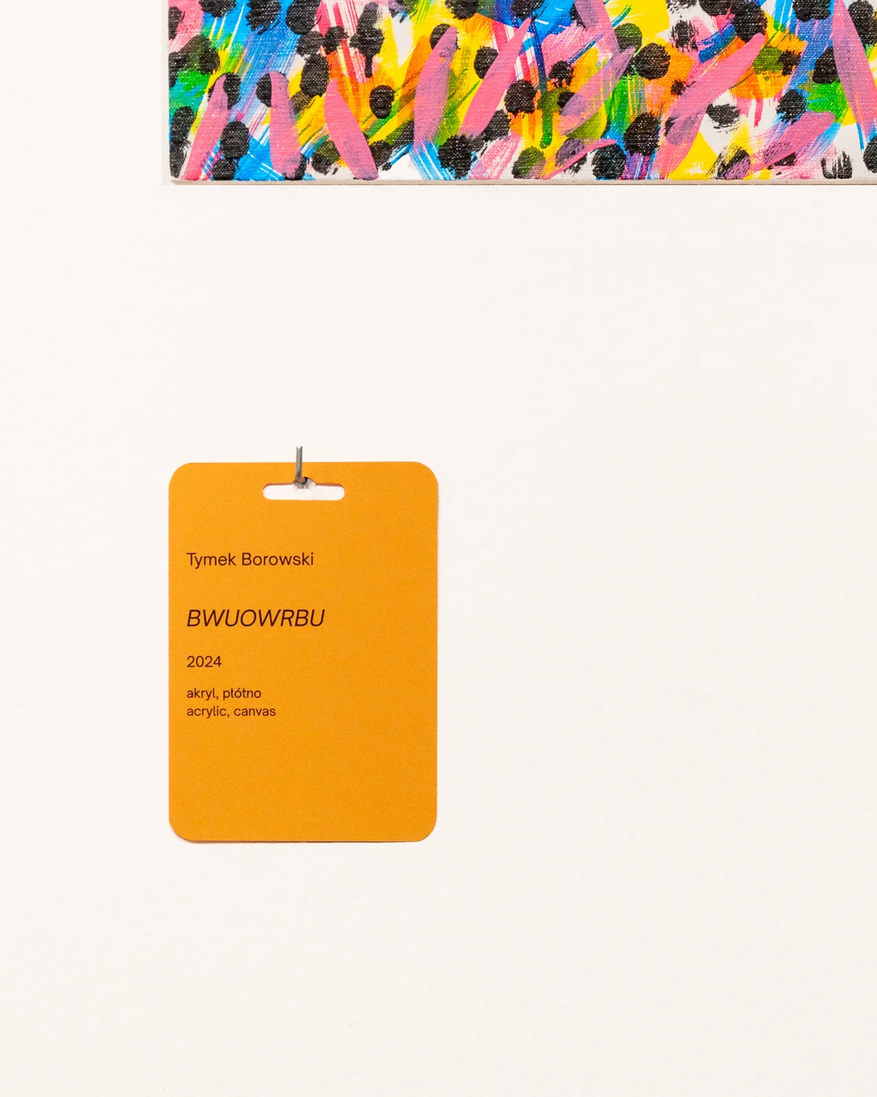 Zbliżenie na żółtą etykietę ścienną do pracy Tymka Borowskiego „BWUOWRBU”, z widocznym powyżej fragmentem kolorowego obrazu.