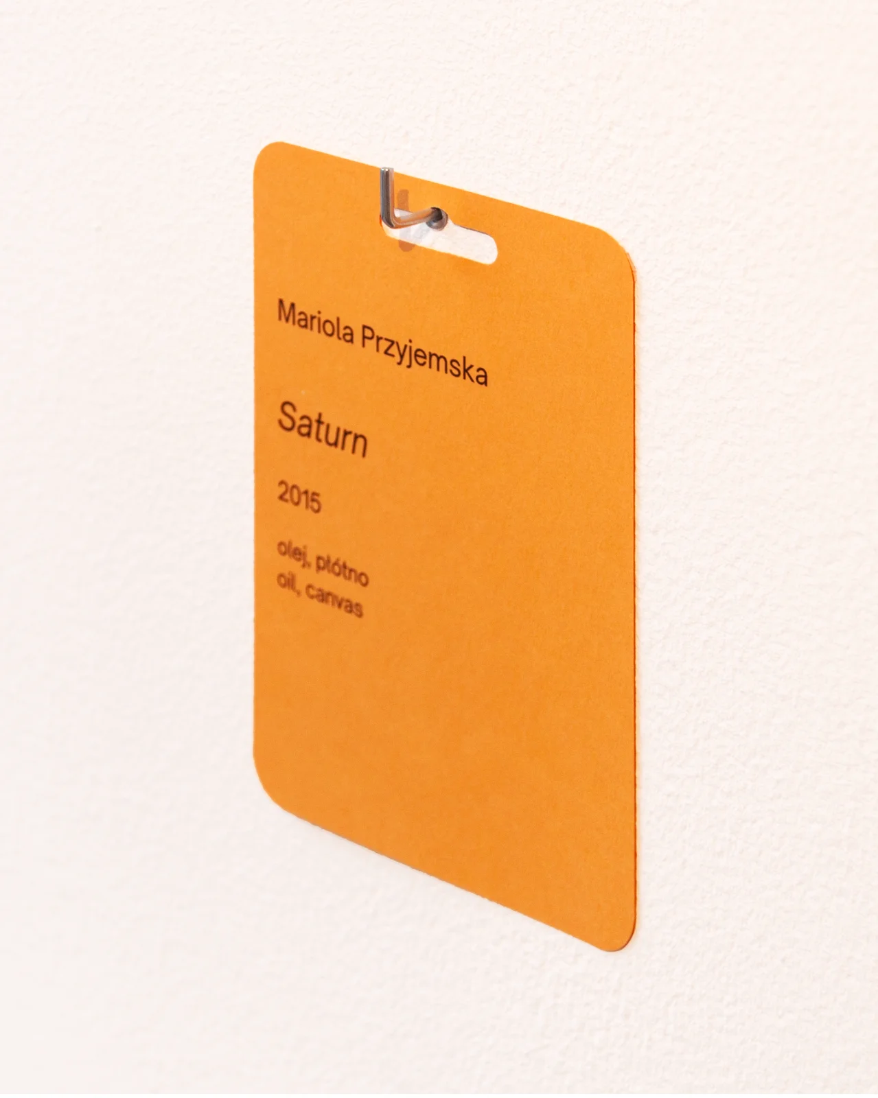Żółta karta identyfikacyjna przypięta do białej ściany, opisująca obraz Marioli Przyjemskiej „Saturn” (2015).