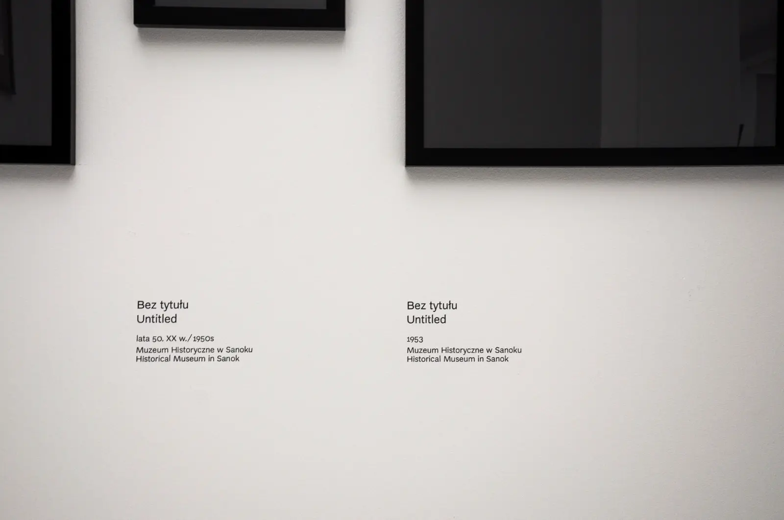 Widok wystawy „Fenomenalny. Zdzisław Beksiński”. Zbliżenie na fragmenty czarnych ram i czarne podpisy na białej ścianie.