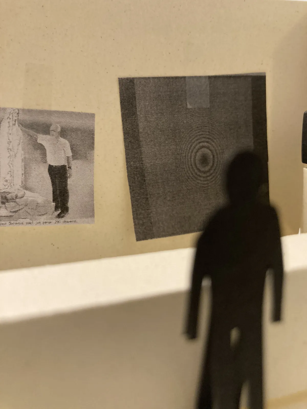 Detal makiety. Na białej ścianie naklejone są dwie małe grafiki: jedna z postacią, druga z abstrakcyjnym motywem kół. Przed nimi stoi czarna, kartonowa sylwetka zwiedzającego.