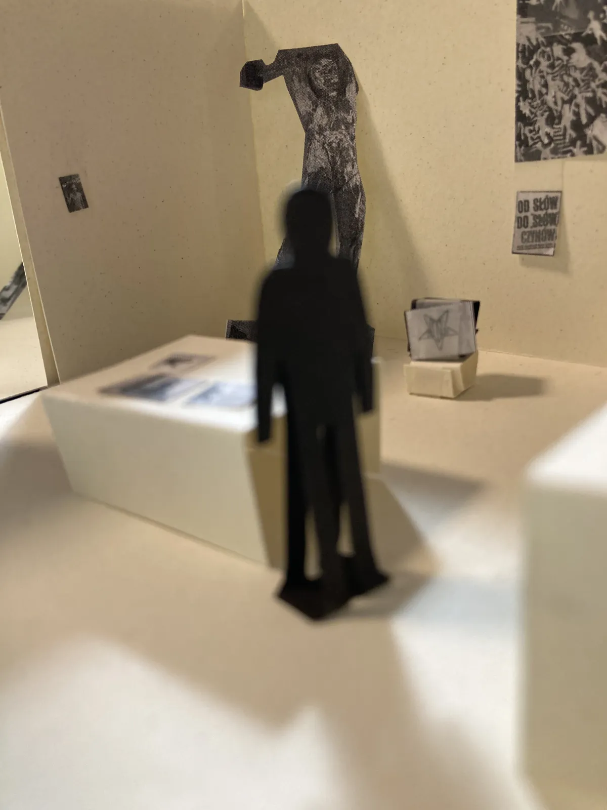 Perspektywiczny widok makiety wystawy. W centralnym punkcie czarna sylwetka zwiedzającego skierowana w stronę miniaturowej, wysokiej rzeźby męskiej postaci.