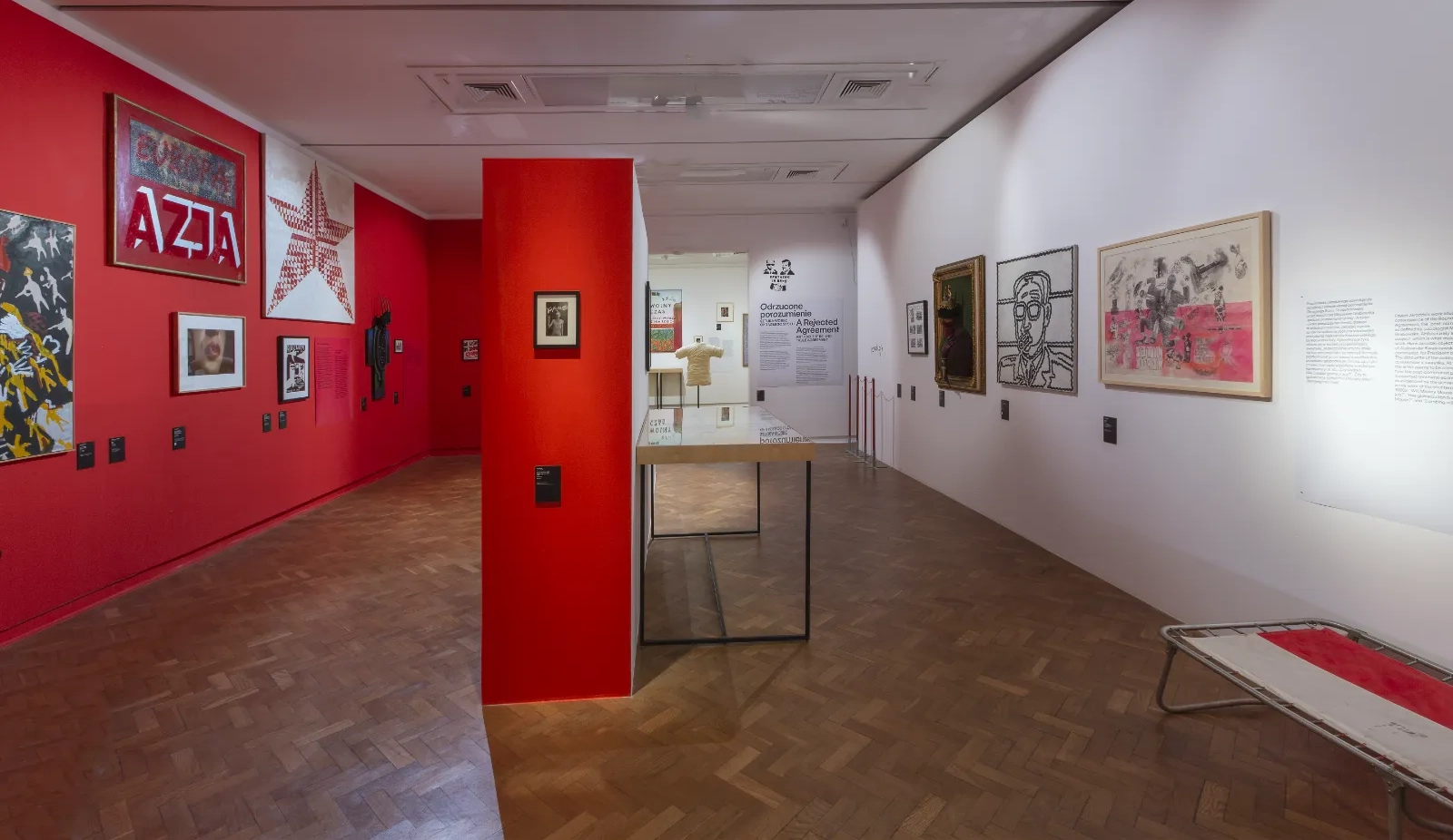 Szerokie ujęcie sali wystawowej. Lewa ściana jest intensywnie czerwona z zawieszonymi obrazami, prawa biała. Na środku stoi czerwony filar i szklana gablota na metalowej konstrukcji.