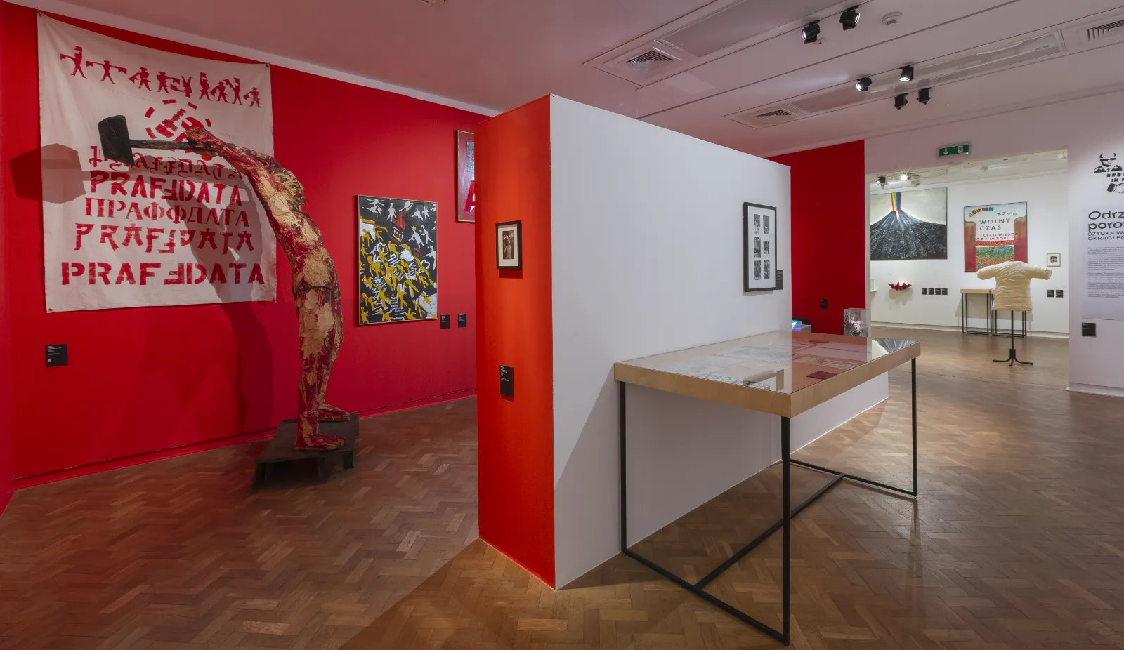 Widok na fragment wystawy z dominującą czerwoną ścianą, na której wisi duży biały transparent. Obok stoi rzeźba przedstawiająca postać trzymającą młot nad głową.