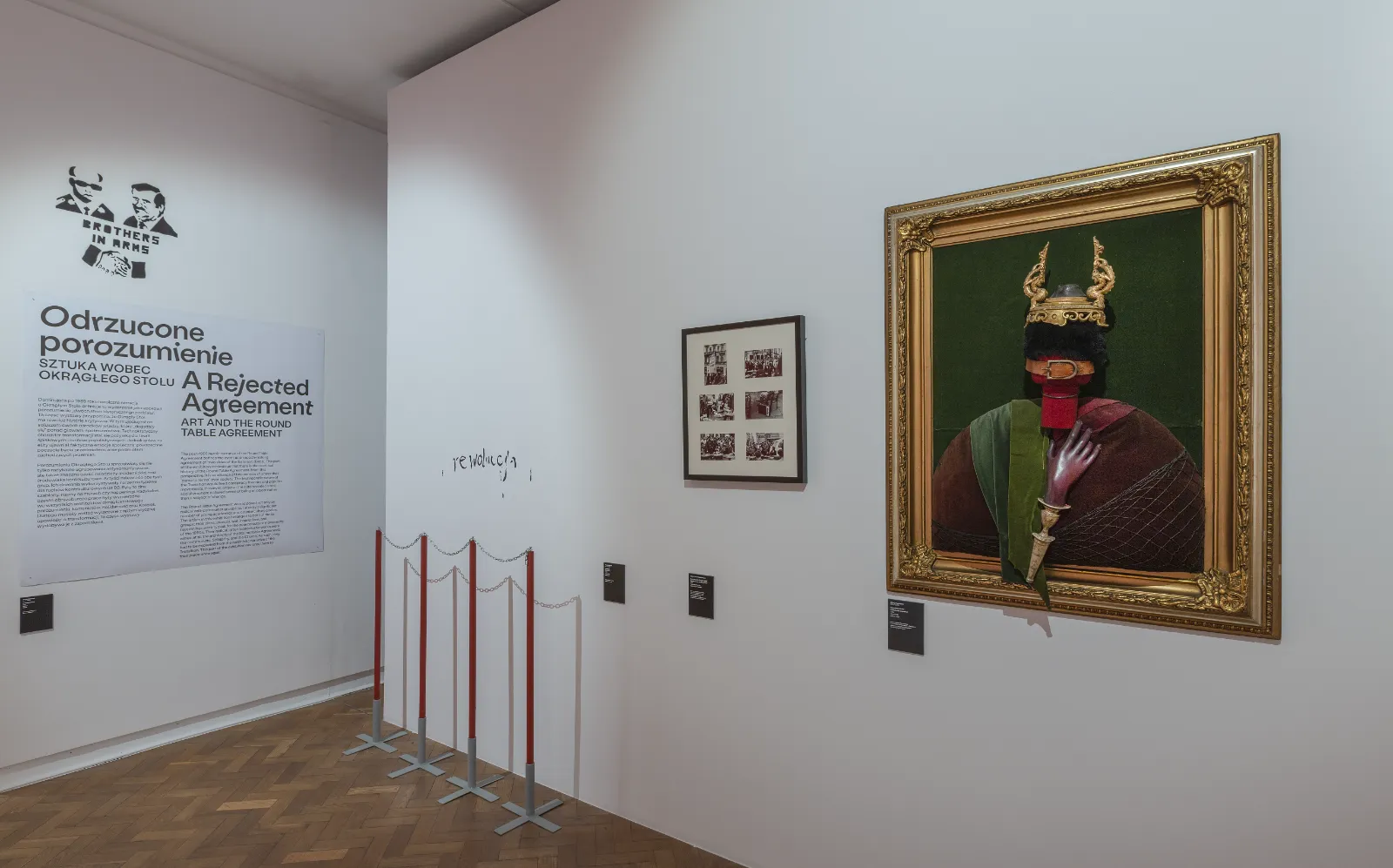 Biała ściana z tekstem kuratorskim „Odrzucone porozumienie”. Obok wisi portret w ozdobnej złotej ramie przedstawiający postać z zasłoniętą twarzą i rogami na głowie.