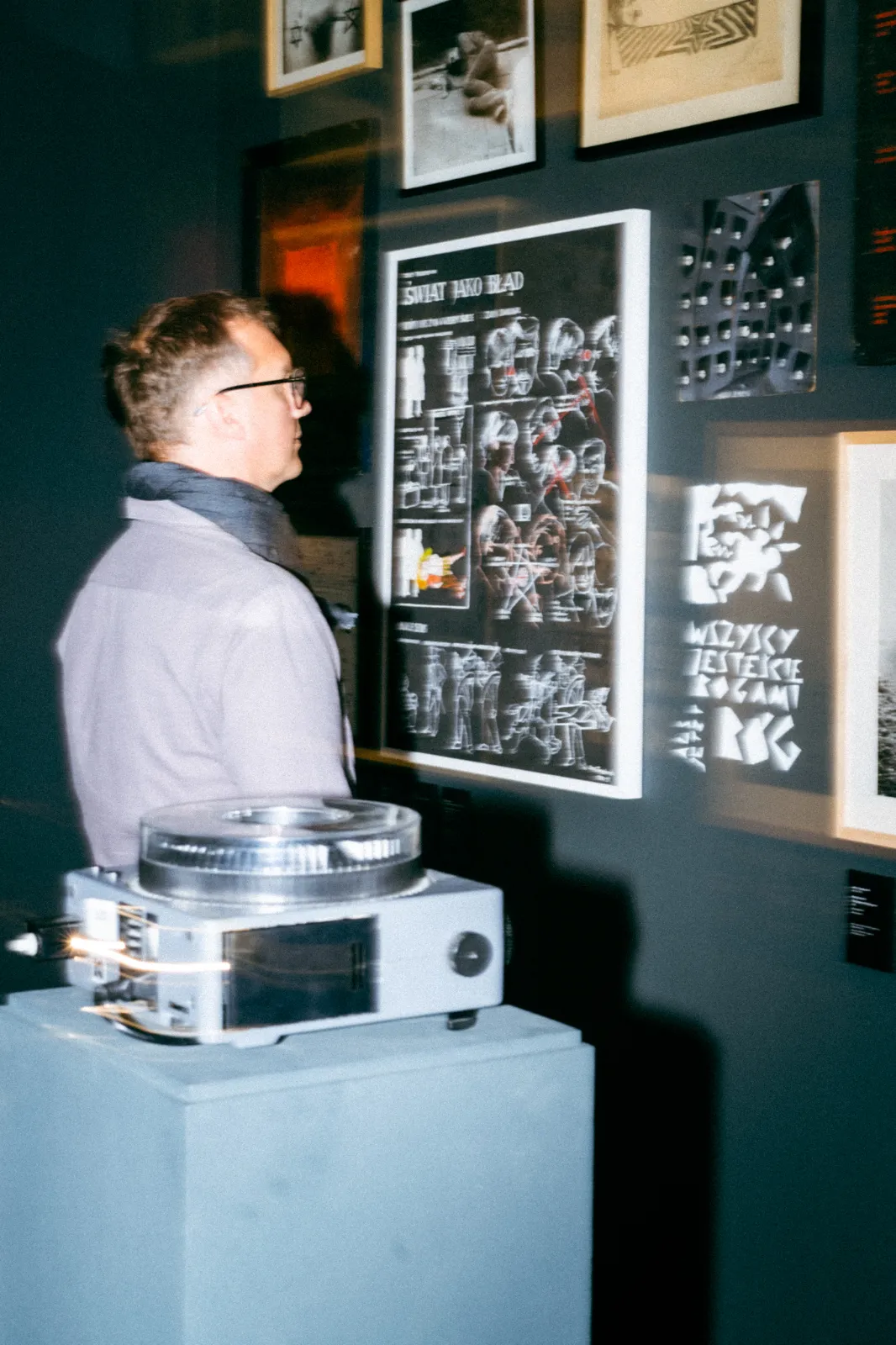 Mężczyzna w okularach ogląda czarną tablicę z grafikami i napisami, m.in. „Świat jako błąd”. Na pierwszym planie, na szarym postumencie, stoi projektor filmowy.