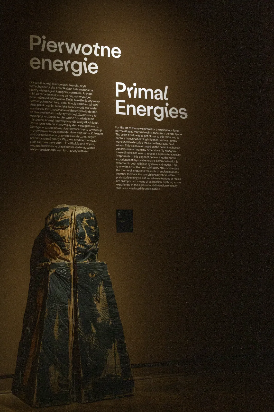 Ciemna sala z podświetlonym tekstem wyklejonym na ścianie „Pierwotne energie”. Poniżej stoi surowa, pionowa rzeźba o teksturze drewna z namalowaną ekspresyjną twarzą.