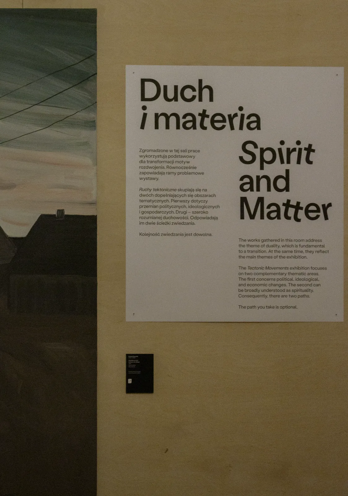Zbliżenie na tablicę informacyjną zatytułowaną „Duch i materia / Spirit and Matter”. Po lewej stronie widać fragment obrazu z mrocznym pejzażem z dachami domów.