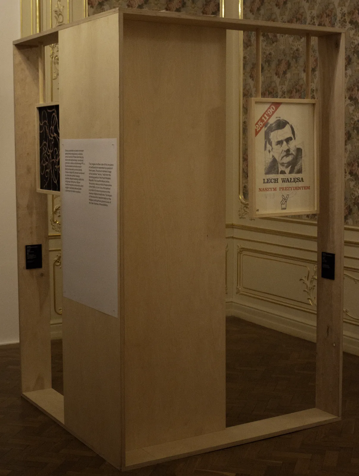 Drewniana konstrukcja w kształcie sześcianu stojąca w sali o wzorzystych tapetach. Na konstrukcji zawieszony jest plakat wyborczy Lecha Wałęsy z 1990 roku.