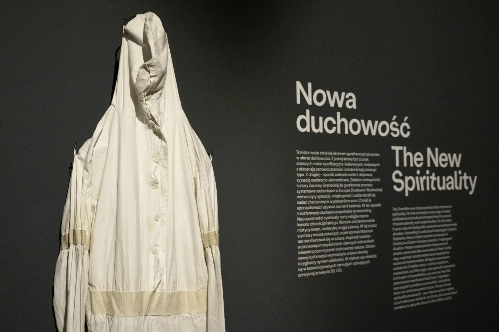 Na ciemnoszarej ścianie zawieszony biały kombinezon z kapturem. Obok widnieje biały napis z tytułem sekcji: „Nowa duchowość”.