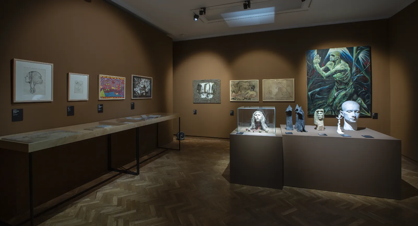 Widok na salę w odcieniach brązu. Na postumentach stoją rzeźby głów, a na ścianie po prawej wisi duży obraz z dynamiczną, zielonkawą postacią w pozie modlitewnej.