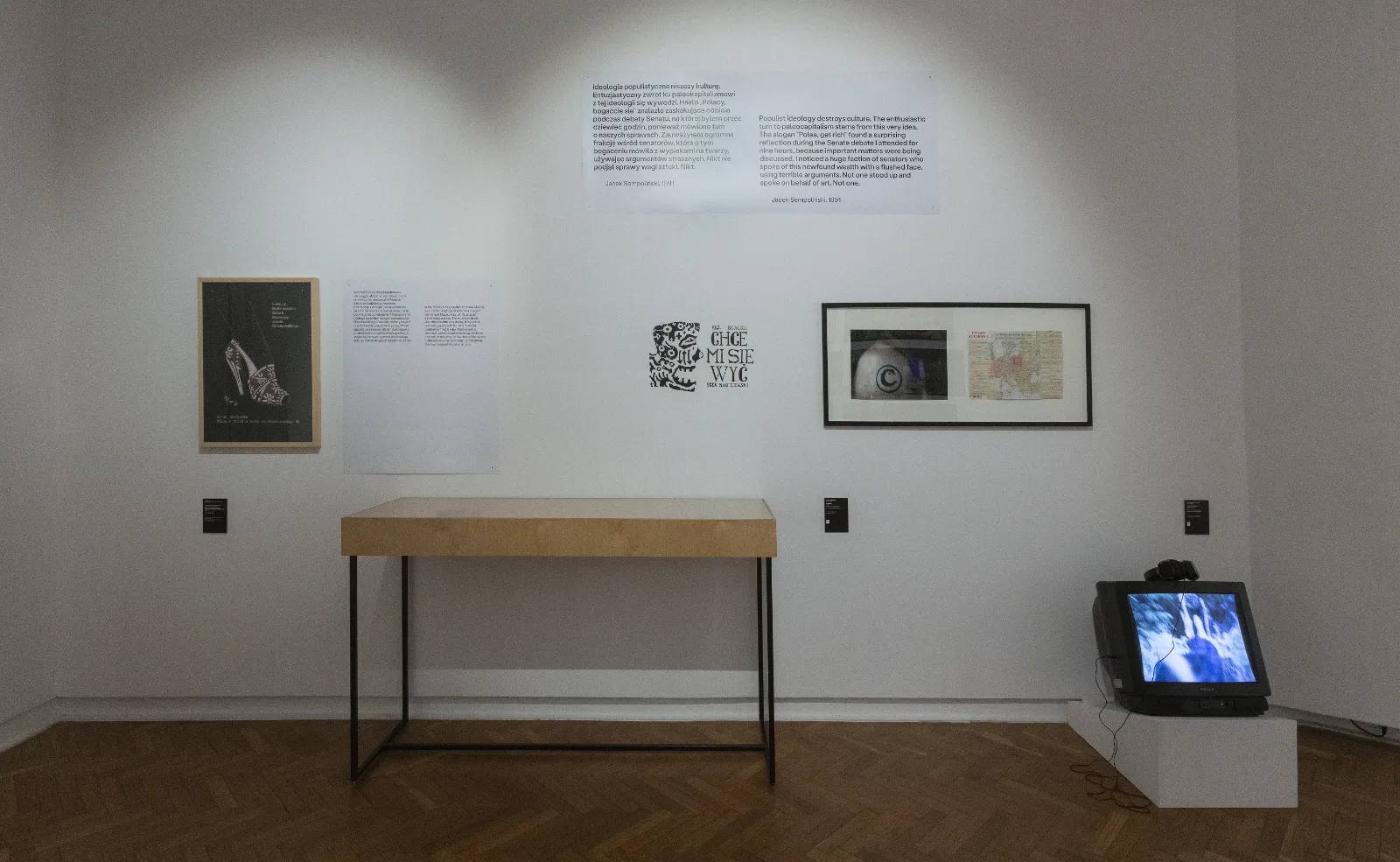 Biała ściana z tekstami i grafikami. Pod ścianą stoi prosty drewniany stolik, a po prawej, na podłodze, stary telewizor kineskopowy wyświetlający niebieskawy obraz.