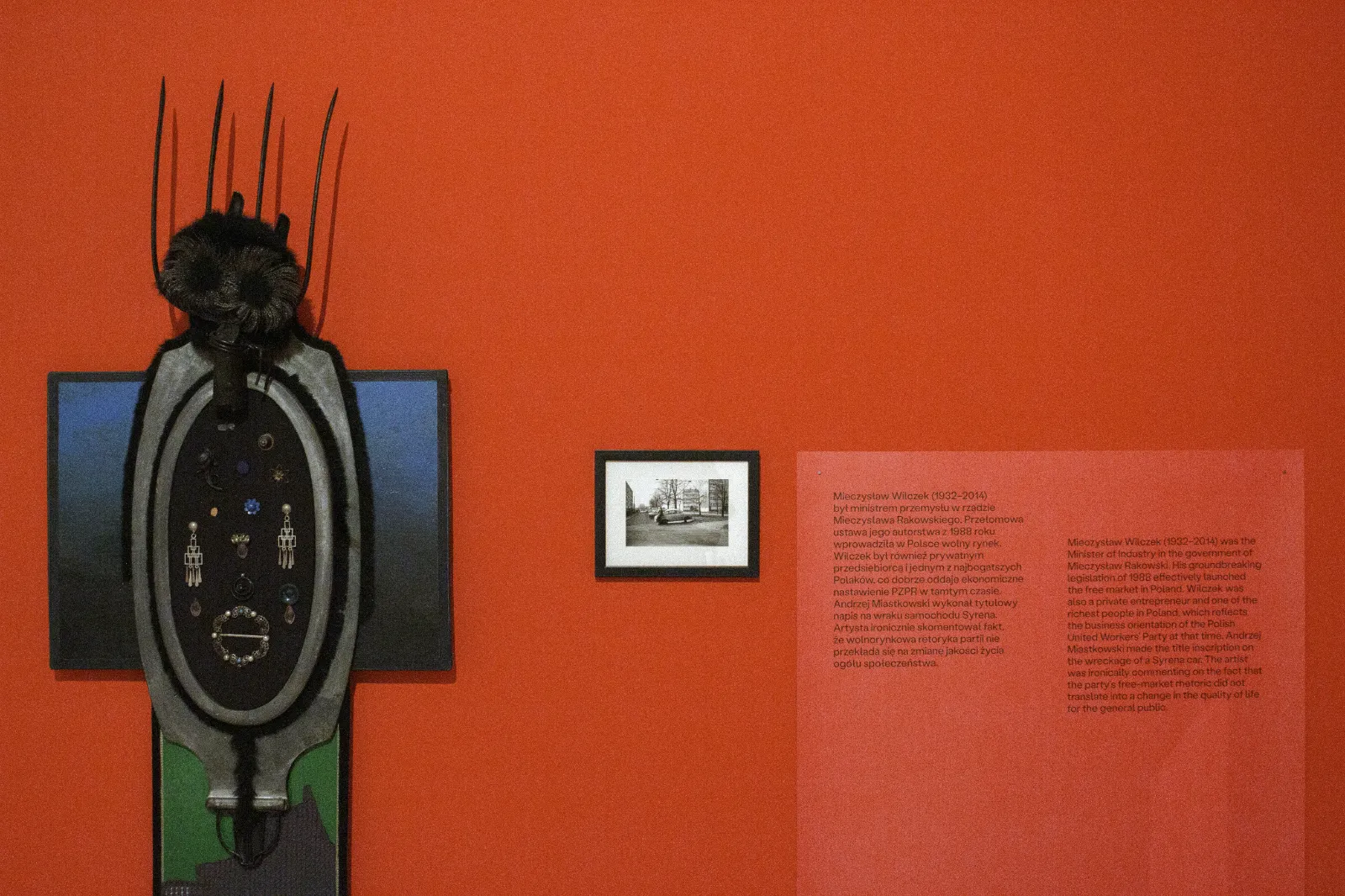 Detal czerwonej ściany z tekstem o Mieczysławie Wilczku. Po lewej wisi asamblaż przypominający maskę lub totem, ozdobiony drobnymi przedmiotami i biżuterią.