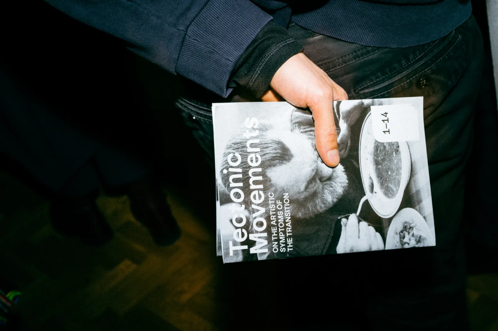 Zbliżenie na dłoń osoby trzymającej broszurę wystawy „Tectonic Movements”. Na okładce znajduje się zdjęcie mężczyzny jedzącego zupę.