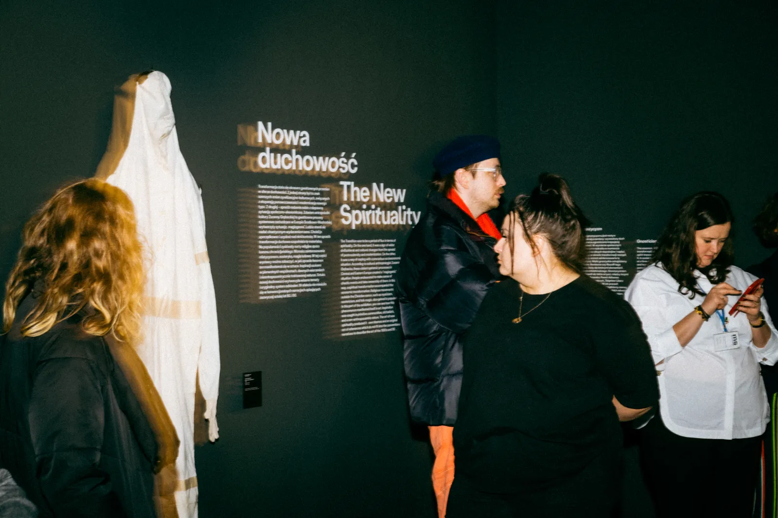 Grupa osób zwiedzających wystawę w sekcji „Nowa duchowość”. W tle widać biały kombinezon i teksty kuratorskie na ciemnej ścianie.