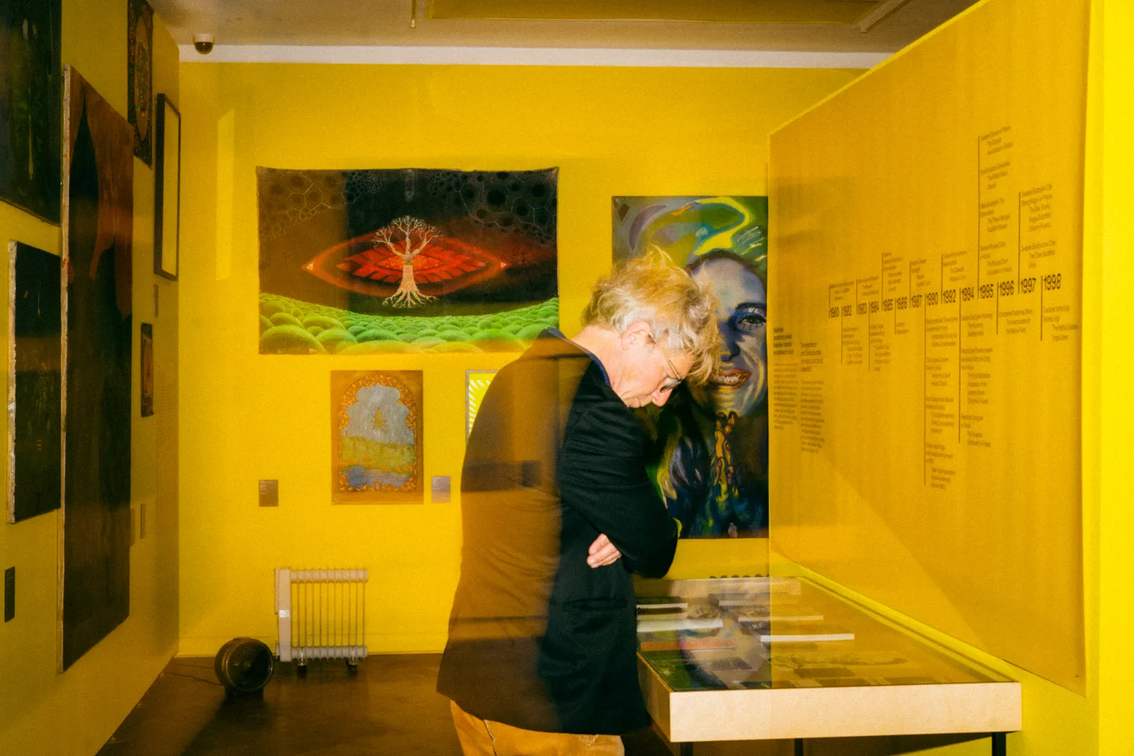 Mężczyzna pochyla się nad szklaną gablotą w sali o żółtych ścianach. W tle widoczne są kolorowe obrazy i tablica z osią czasu transformacji.