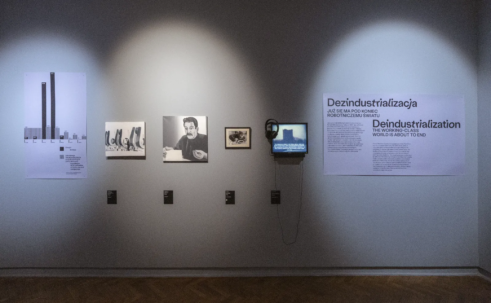 Szara ściana z sekcją „Dezindustrializacja”. Widać na niej wykres słupkowy, małe obrazy (w tym portret robotnika) oraz monitor z wyświetlanym filmem.
