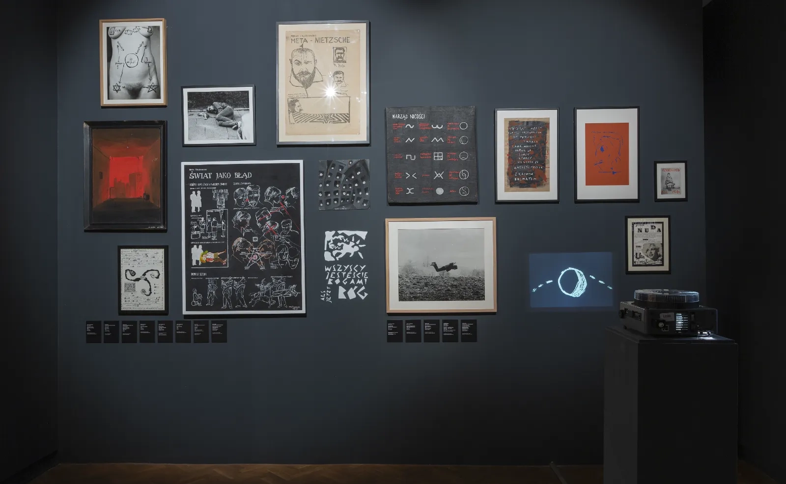 Ciemna ściana gęsto wypełniona różnorodnymi pracami: grafikami, zdjęciami i rysunkiem z napisem „Meta-Nietzsche”. Obok stoi projektor na postumencie.