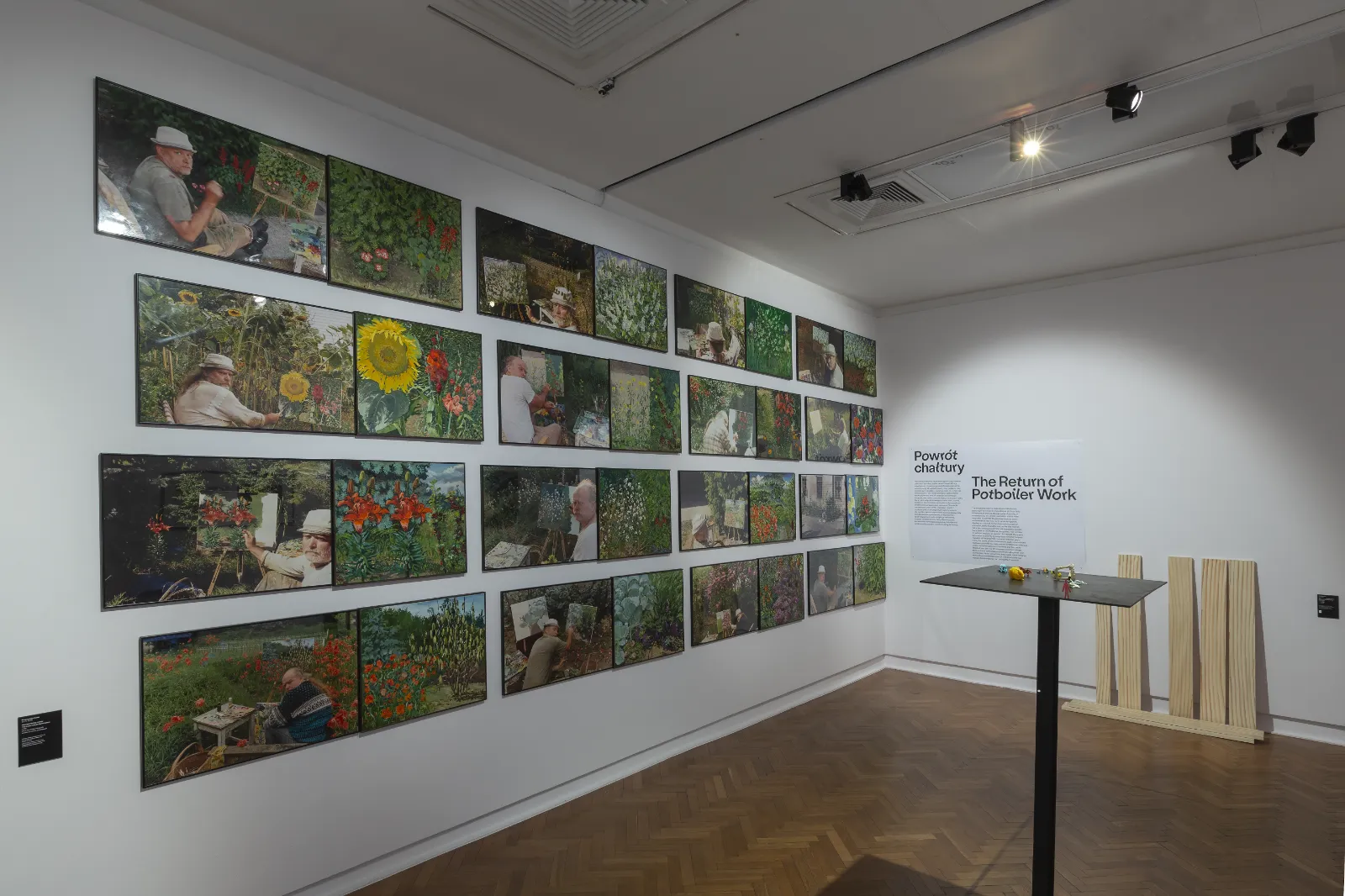 Ściana w białej sali pokryta siatką kilkunastu małych obrazów przedstawiających tego samego mężczyznę malującego w plenerze. Obok tekst „Powrót chałtury”.