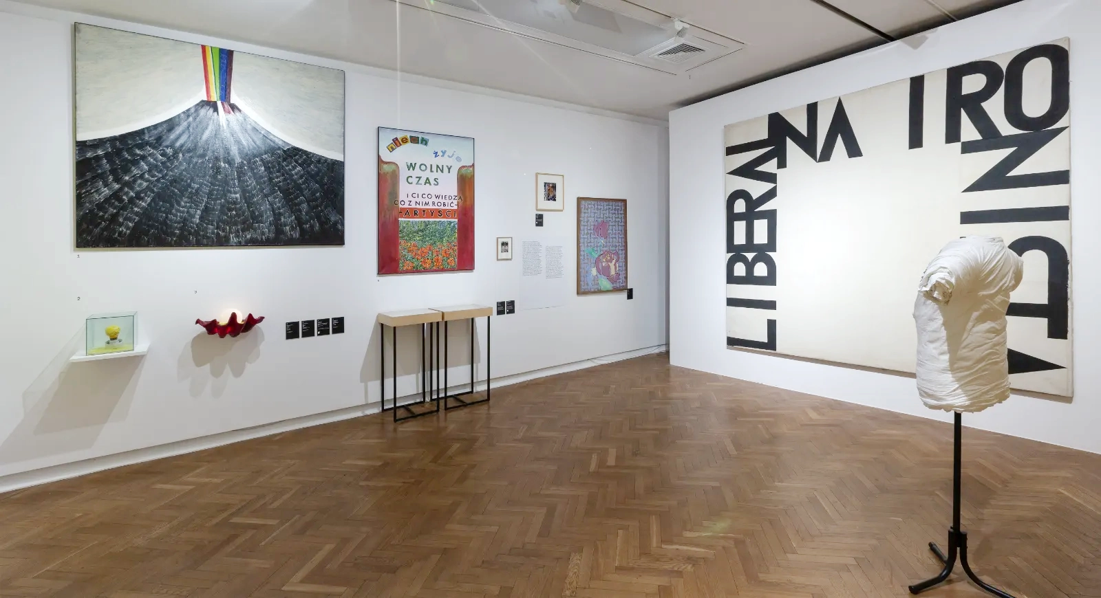 Widok sali z dużym obrazem „Liberalna ironista” (wielkie czarne litery na białym tle). Obok wisi płótno z tęczowym wulkanem, a przed nimi stoi biała koszulka na stojaku.