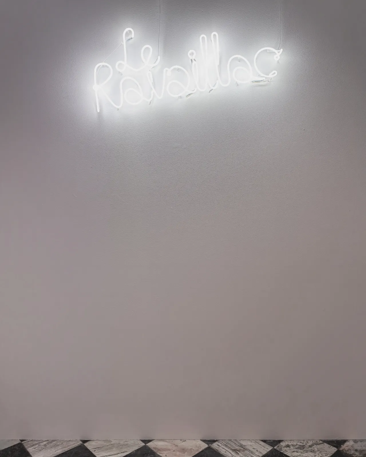 Świecący biały neon z napisem „Le Ravaillac” zamontowany na białej ścianie galerii.