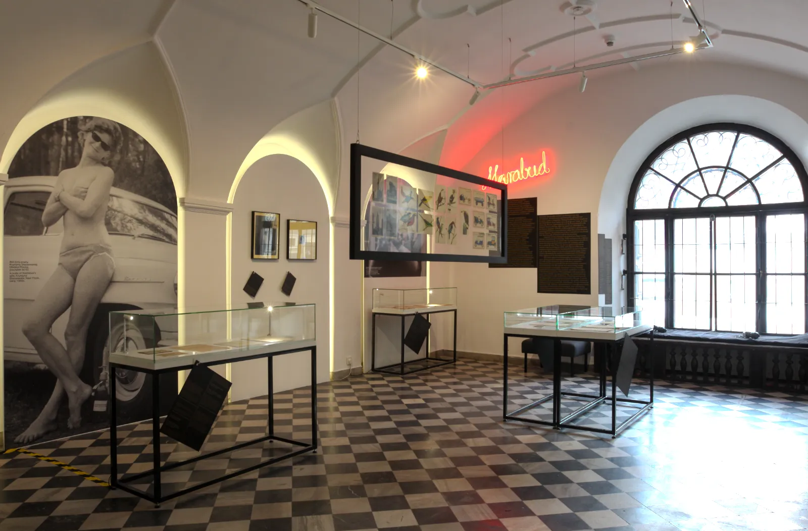 Szerokie ujęcie sali z szachownicą na podłodze, czerwonym neonem „Marabud” i zdjęciem żony artysty przy samochodzie.