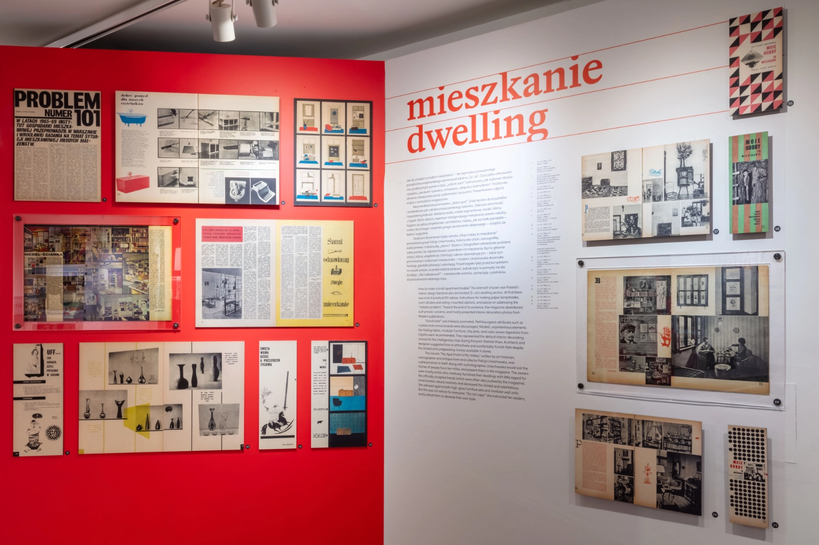 Część wystawy „mieszkanie / dwelling”: rozkładówki o urządzaniu wnętrz na czerwonej ścianie oraz biała ściana z dwujęzycznym tekstem i czerwonym tytułem.