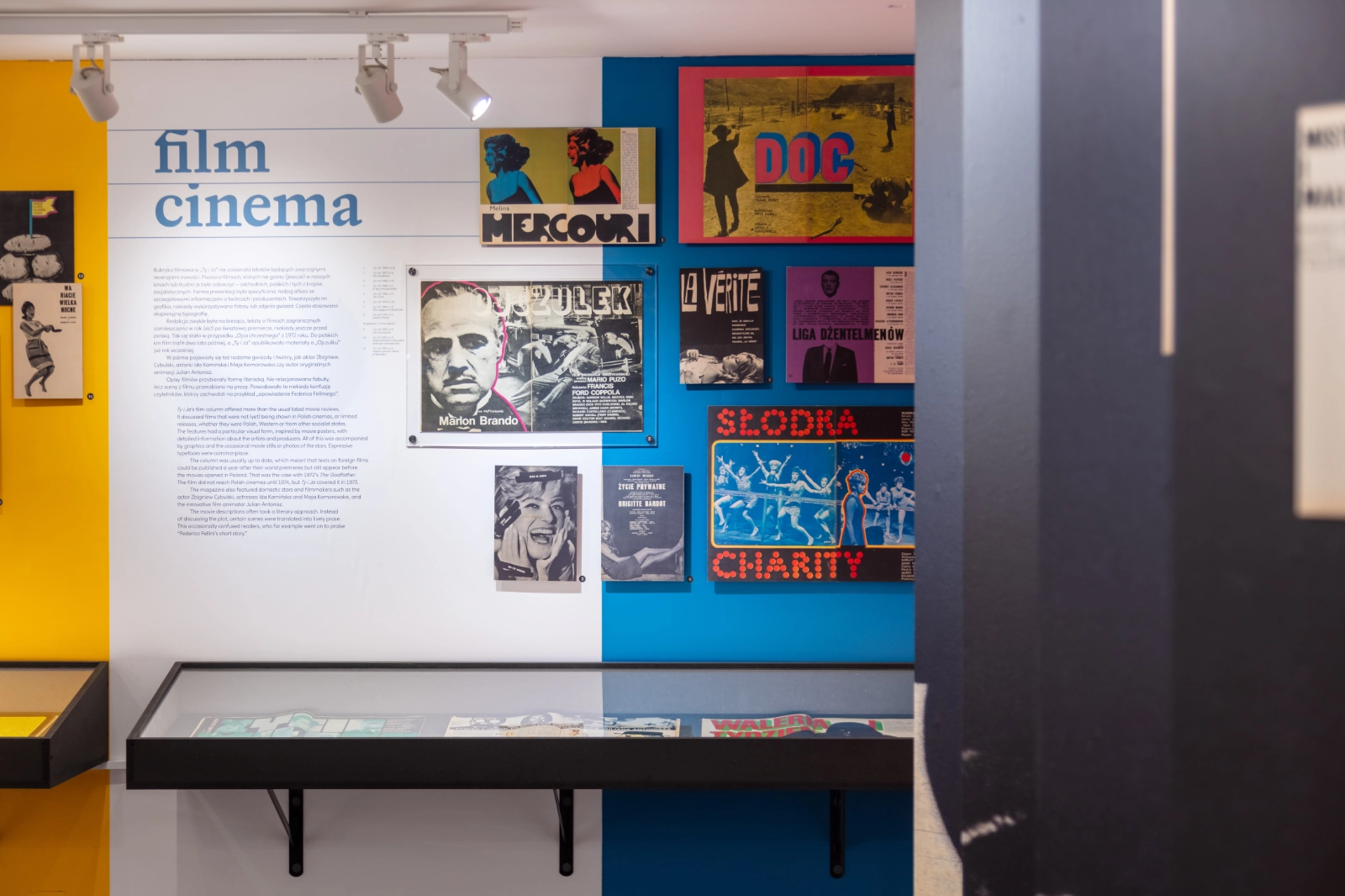 Część wystawy „film / cinema”: biała ściana z niebieskim tytułem i dwujęzycznym tekstem obok niebieskiej ściany z polskimi plakatami filmowymi i stronami magazynu, poniżej gablota.