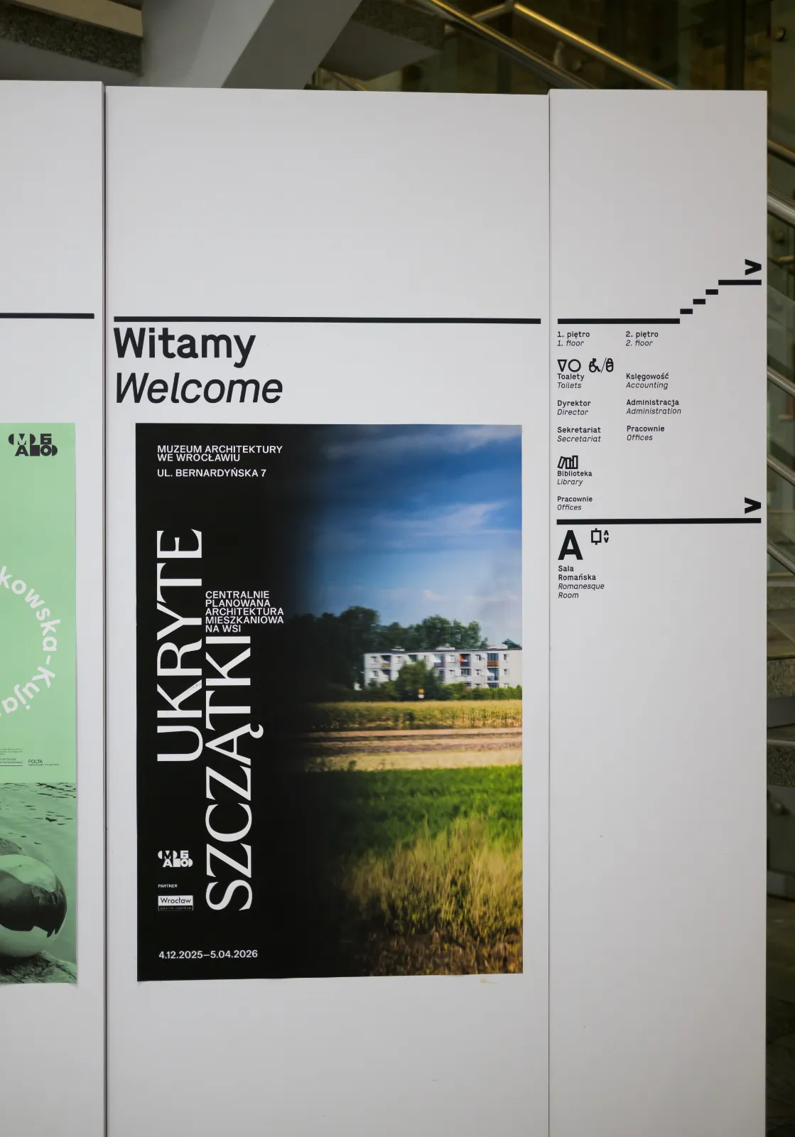 Plakat wystawy na tablicy informacyjnej: tytuł po lewej stronie, fotografia przedstawiająca blok pośród pól po prawej.